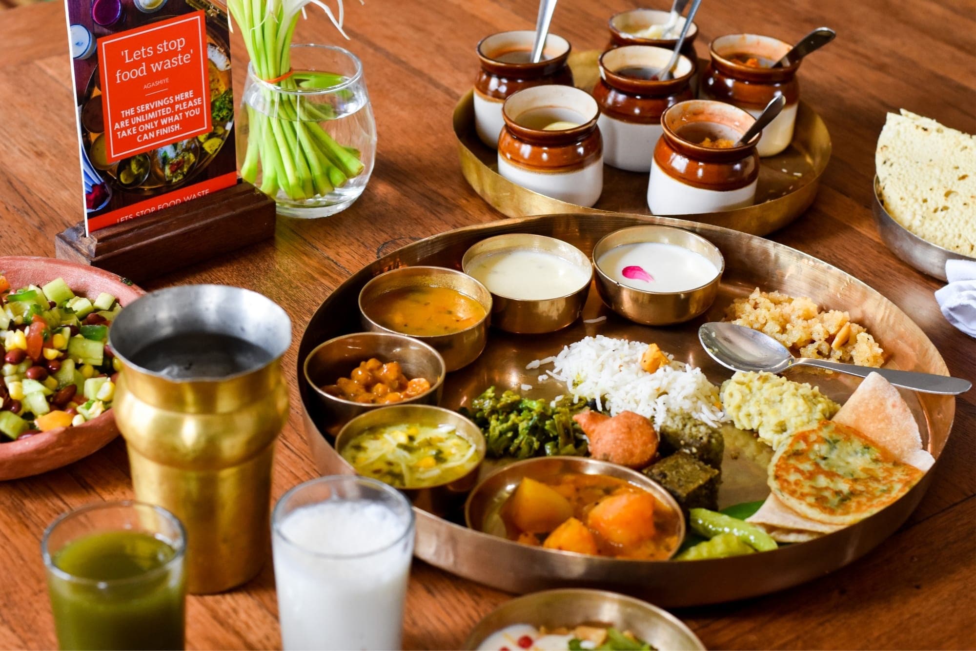 A Gujarati thali on a brass plate with small bowls of dal, kadhi, shaak, rice and rotli on a wooden table