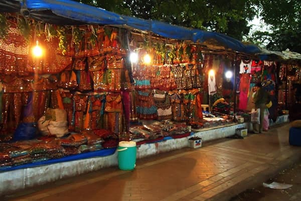 A row of lit market stalls hung with embroidered and mirror-work garments and bags in the evening