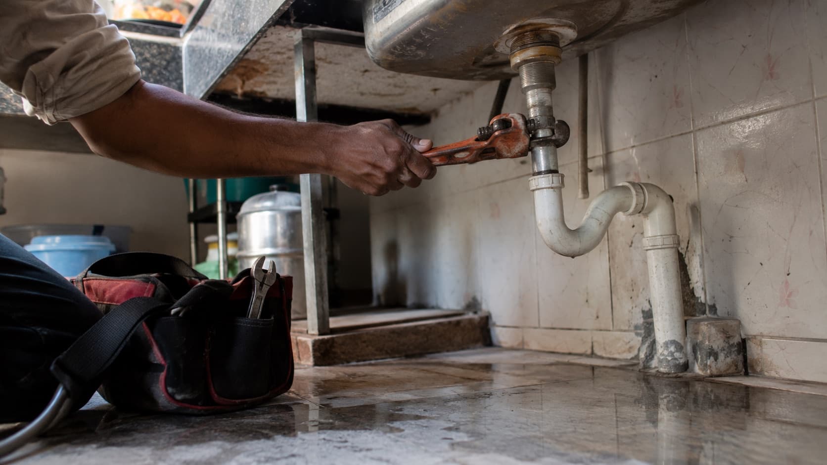 A hand tightening a pipe joint with a wrench beneath a steel kitchen sink, tool bag on the floor