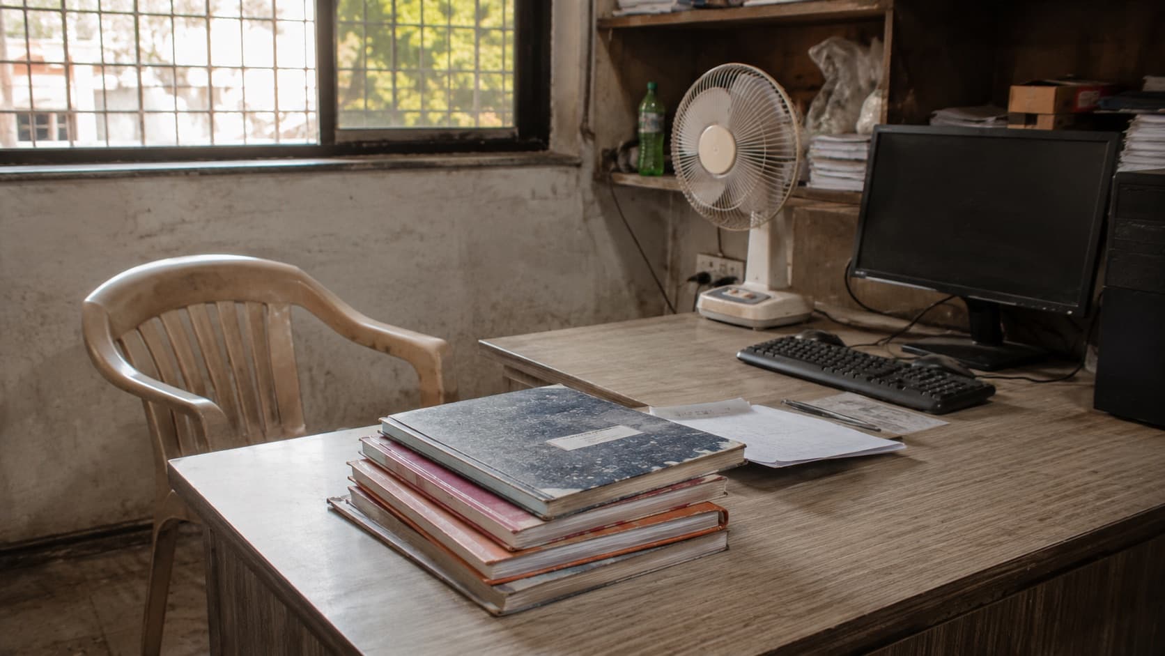 A desk in a small shop back office with a computer, stacked ledger books and an empty plastic chair